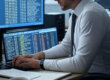 A realistic office scene showing a cybersecurity analyst reviewing Google Drive, Dropbox, and OneDrive logs on dual monitors. One screen displays a cloud activity audit log with timestamps and IP addresses, the other shows a data breach report. The workspace includes a notepad, coffee, and sticky notes labeled “Cloud Forensics.”