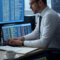 A realistic office scene showing a cybersecurity analyst reviewing Google Drive, Dropbox, and OneDrive logs on dual monitors. One screen displays a cloud activity audit log with timestamps and IP addresses, the other shows a data breach report. The workspace includes a notepad, coffee, and sticky notes labeled “Cloud Forensics.”