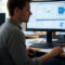 A realistic photo of a small business IT technician analyzing network traffic logs on a dual-monitor setup in a modest office. One screen shows Wireshark or similar forensic software, the other has a company network diagram. The setting is professional yet casual, with papers and coffee cups nearby.